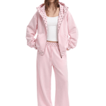 A model wearing a light pink fleece tracksuit with a matching zip-up hoodie, white tank top, and white sandals, set against a plain white background.