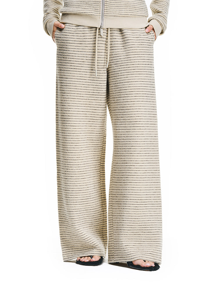 A front view of a model wearing cream-colored, wide-leg textured lounge pants with a matching zip-up hoodie, featuring a relaxed fit and drawstring waist.