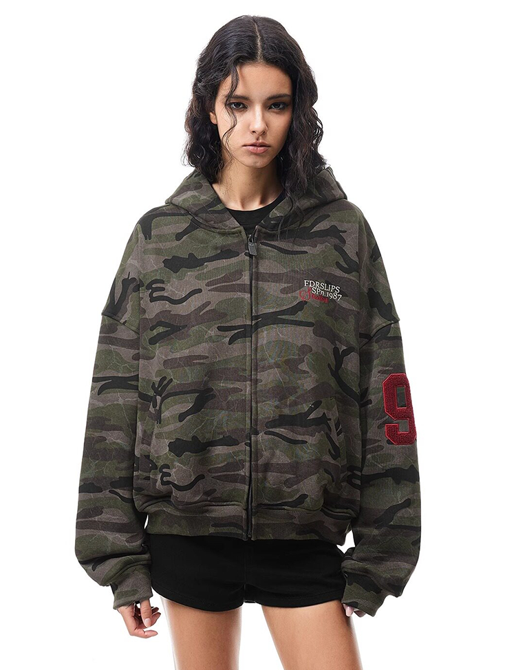 Front view of model wearing camo hoodie with small chest embroidery and red number print on sleeve.