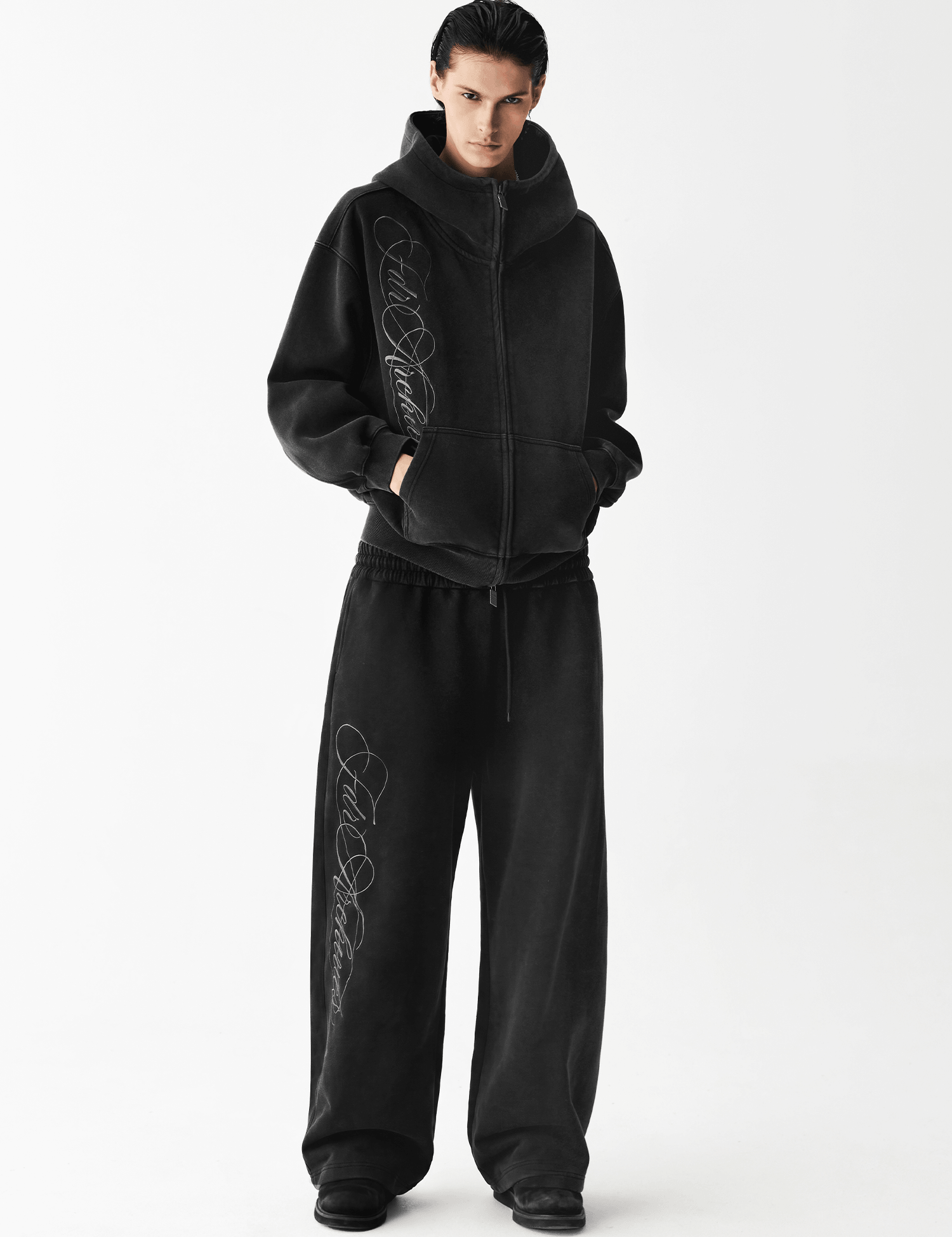 Full body shot of a male model wearing an oversized washed black embroidered zip hoodie and matching wide-leg pants.