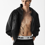 Close-up front view of a male model with an unzipped washed black embroidered hoodie over a bare chest.