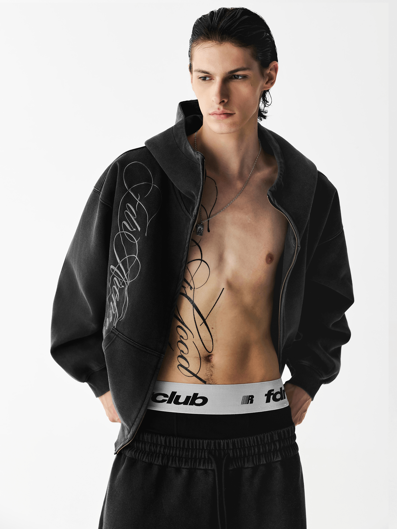 Close-up front view of a male model with an unzipped washed black embroidered hoodie over a bare chest.