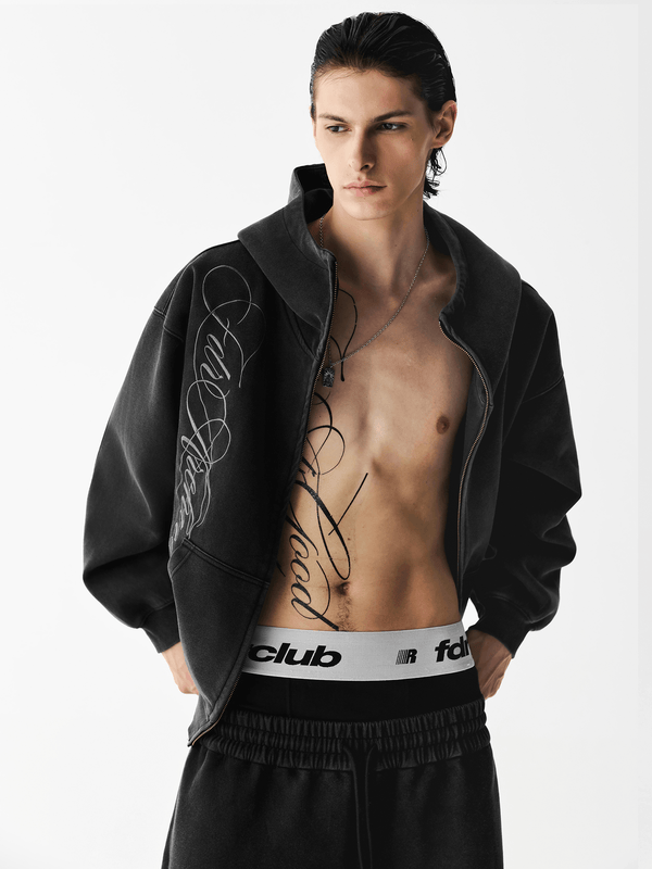 Close-up front view of a male model with an unzipped washed black embroidered hoodie over a bare chest.