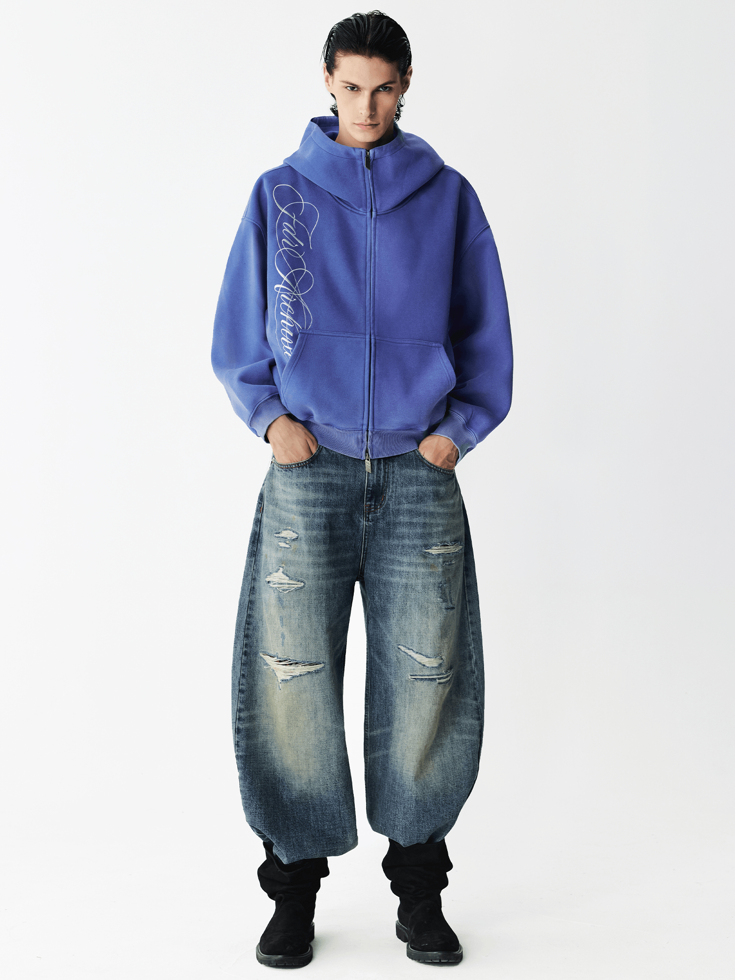 Full body shot of a male model wearing a washed blue embroidered zip hoodie with wide-leg distressed jeans.