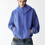 Half-body view of a male model wearing a fully zipped high-neck washed blue embroidered hoodie with hands in pockets.