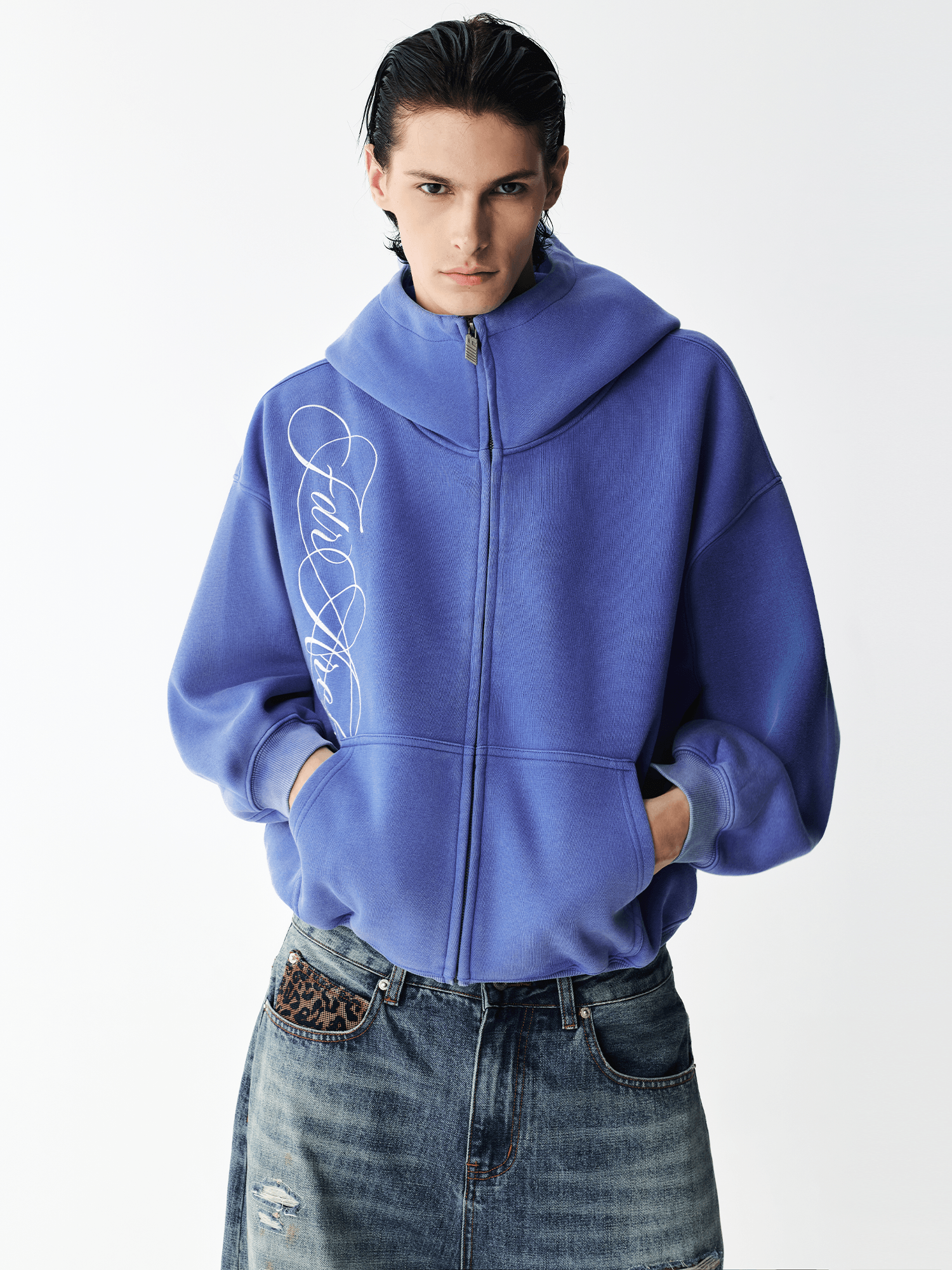 Half-body view of a male model wearing a fully zipped high-neck washed blue embroidered hoodie with hands in pockets.