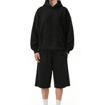 Full-body front view of a model wearing a solid black hoodie and matching shorts, paired with white socks and black shoes (featuring a checkbox in the upper-left corner).