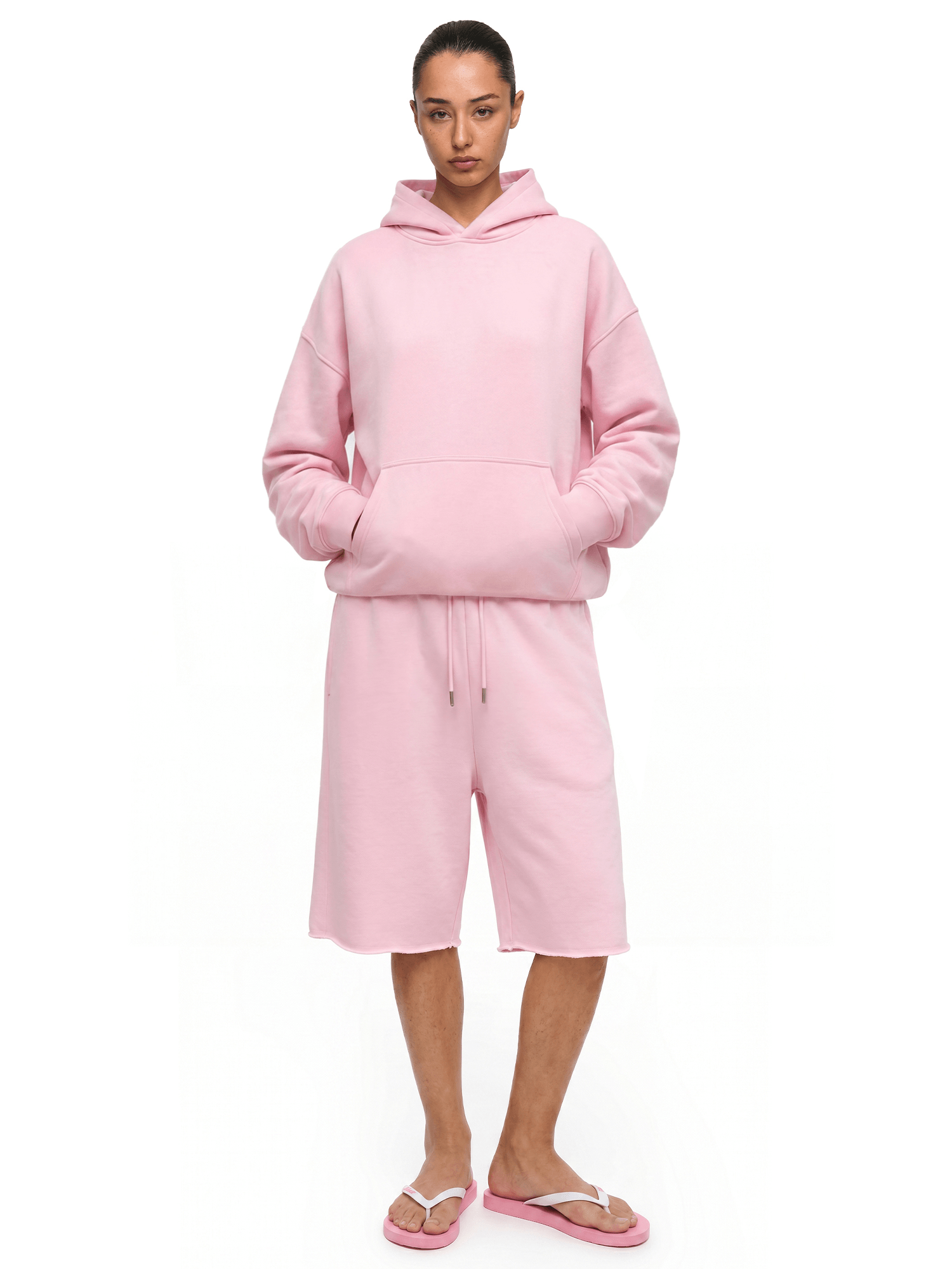 Full-body front view of a model wearing a light pink hoodie and matching loose over-the-knee shorts, paired with pink sandals.