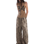 Three-quarter view of a model in a brown "24" graphic sleeveless top and oversized leopard print wide-leg pants.