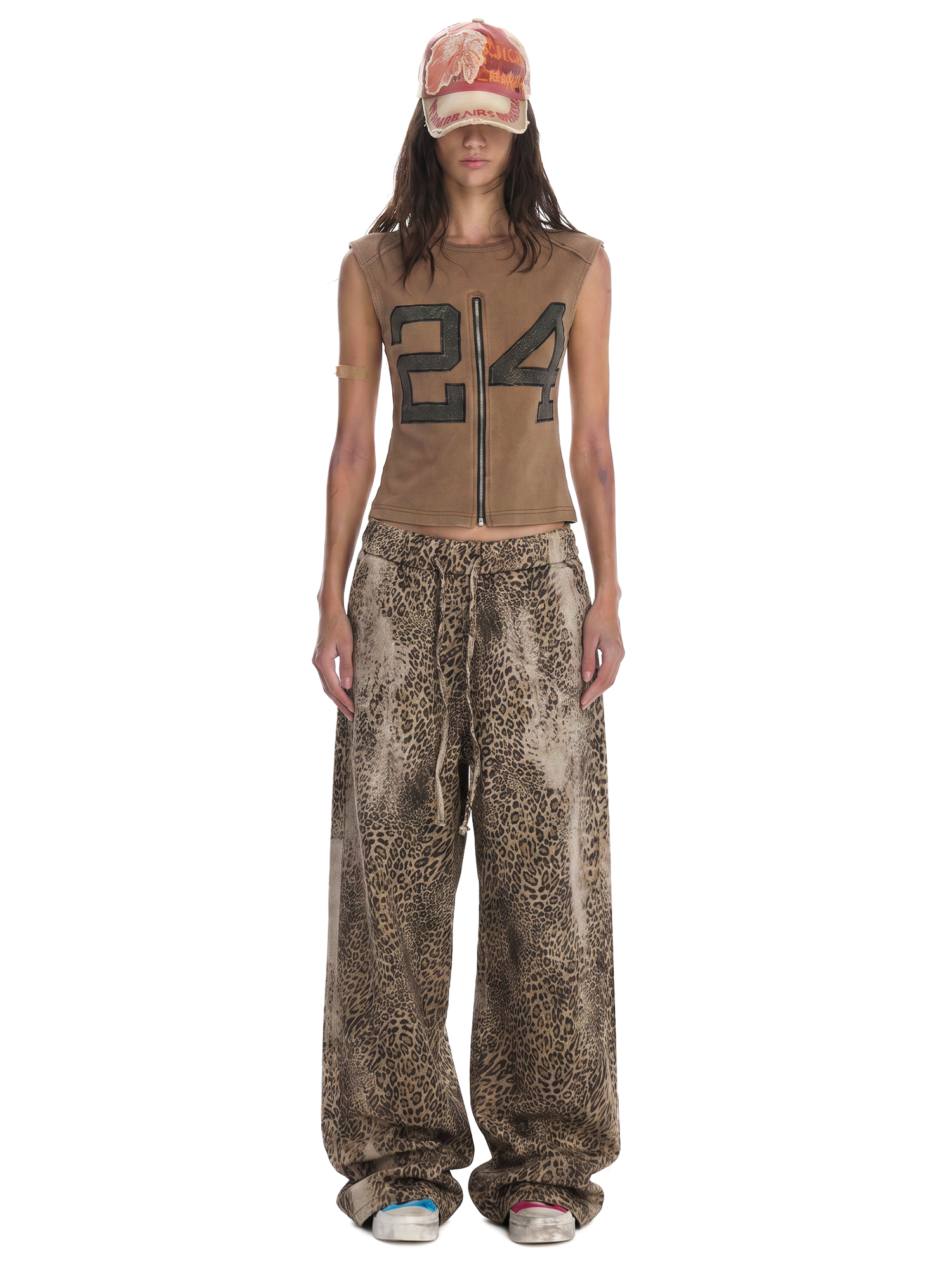 Full-length front view of a model wearing a brown sleeveless graphic zip-up top and oversized leopard print drawstring trousers.