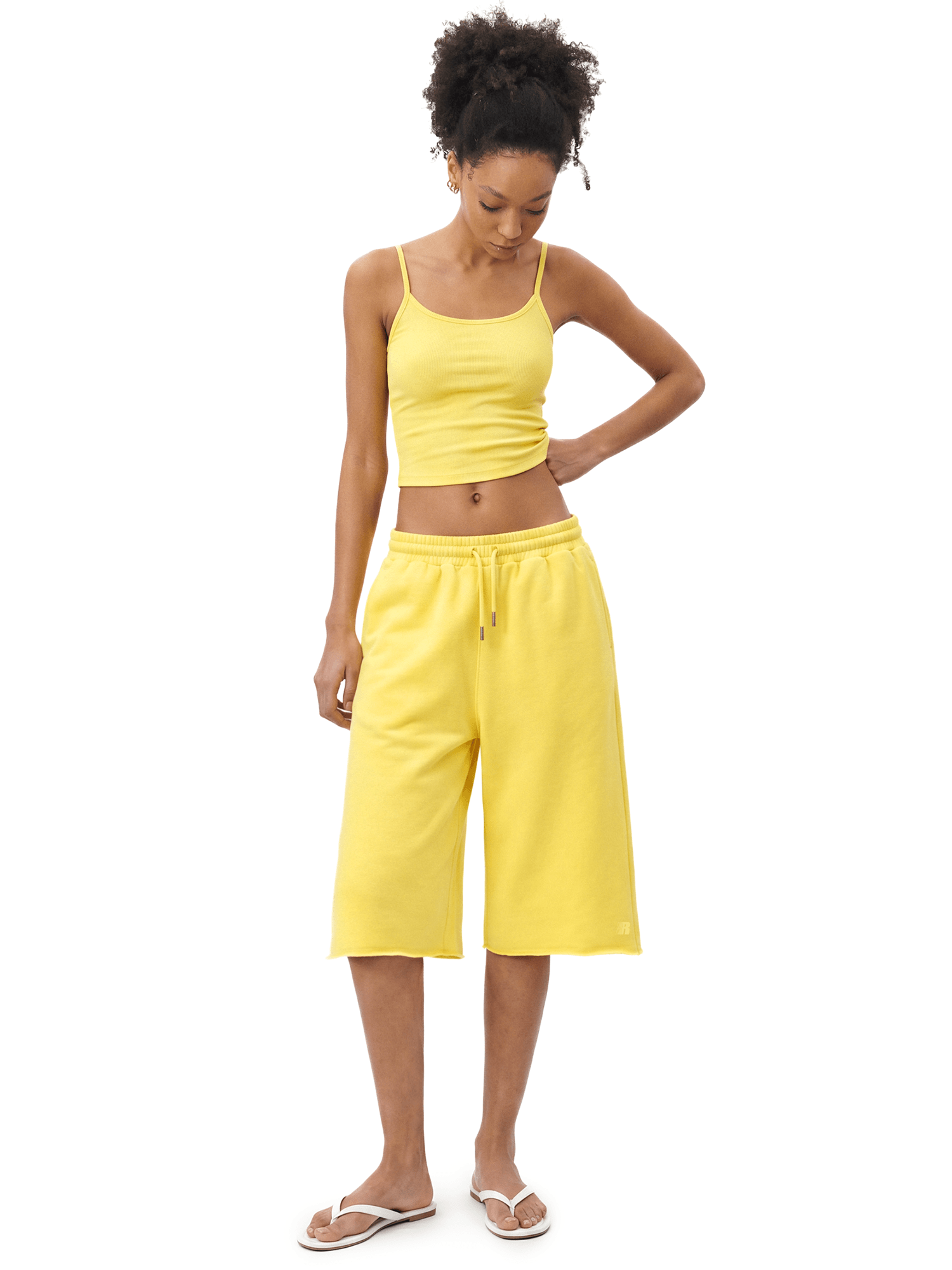 Full-body front view of a female model wearing a bright yellow spaghetti-strap camisole cropped top and matching bright yellow knee-length loose shorts, looking down with one hand on her hip.