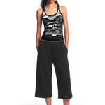 Full-body front view of a female model wearing a black tank top with a graphic print on the front, black wide-leg cropped pants, and black flip-flops, standing with her hands in her pockets against a plain white background.