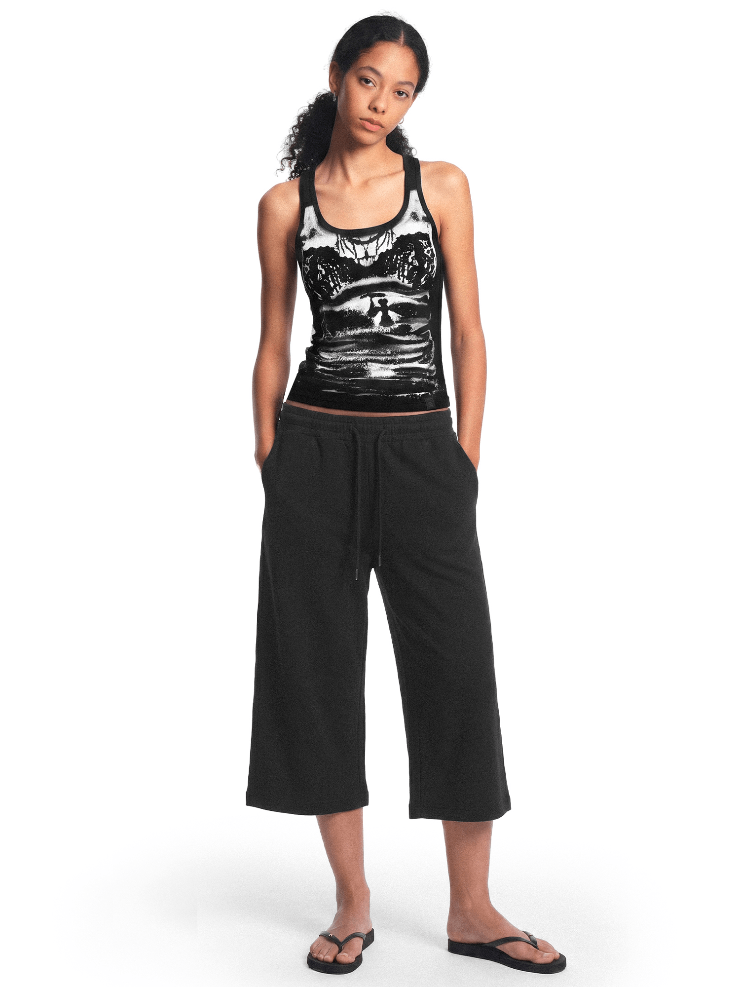 Full-body front view of a female model wearing a black tank top with a graphic print on the front, black wide-leg cropped pants, and black flip-flops, standing with her hands in her pockets against a plain white background.
