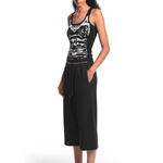 Full-body view of a model from a slight angle wearing the black graphic tank top, black cropped pants, and sandals.
