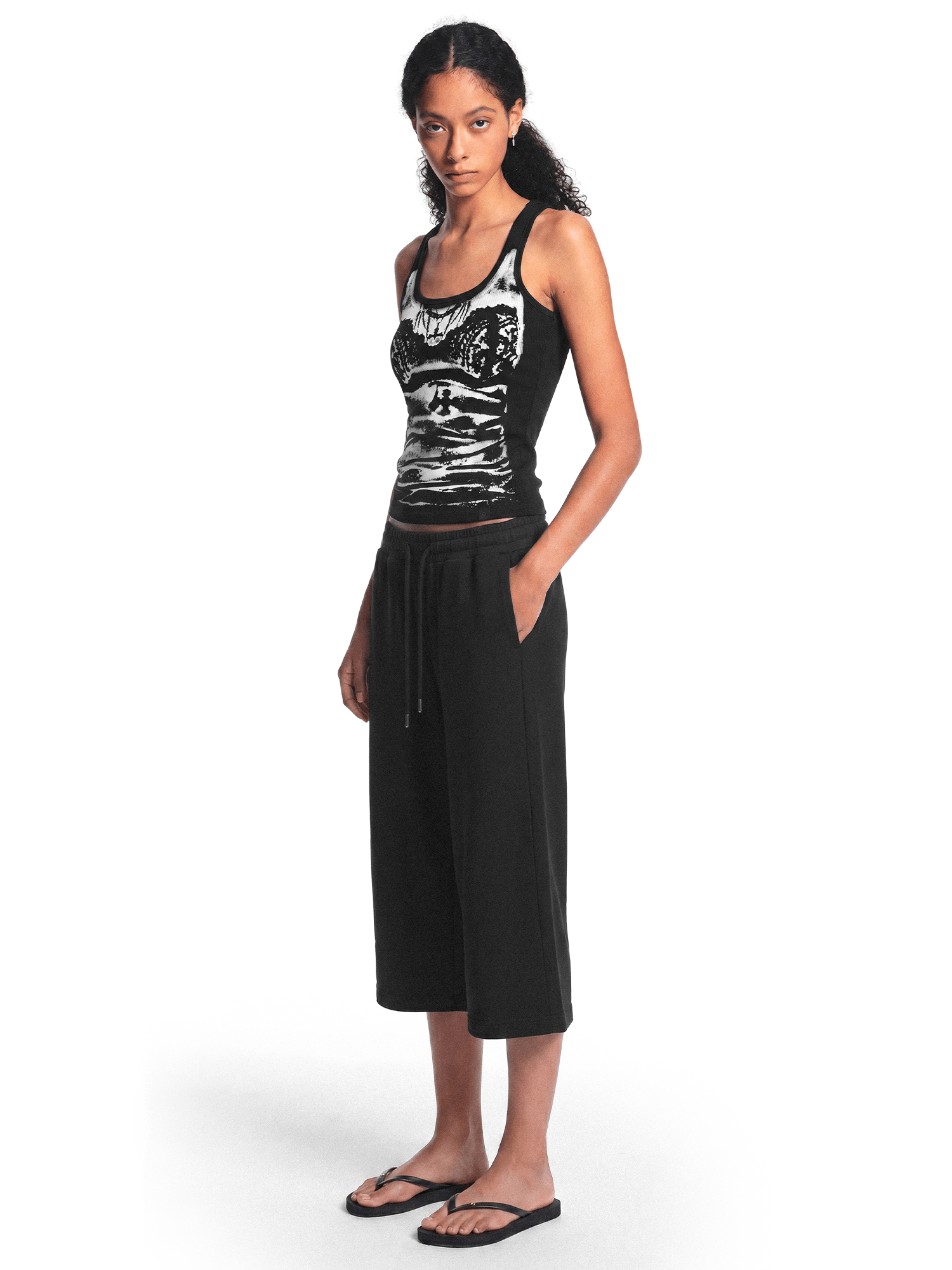 Full-body view of a model from a slight angle wearing the black graphic tank top, black cropped pants, and sandals.