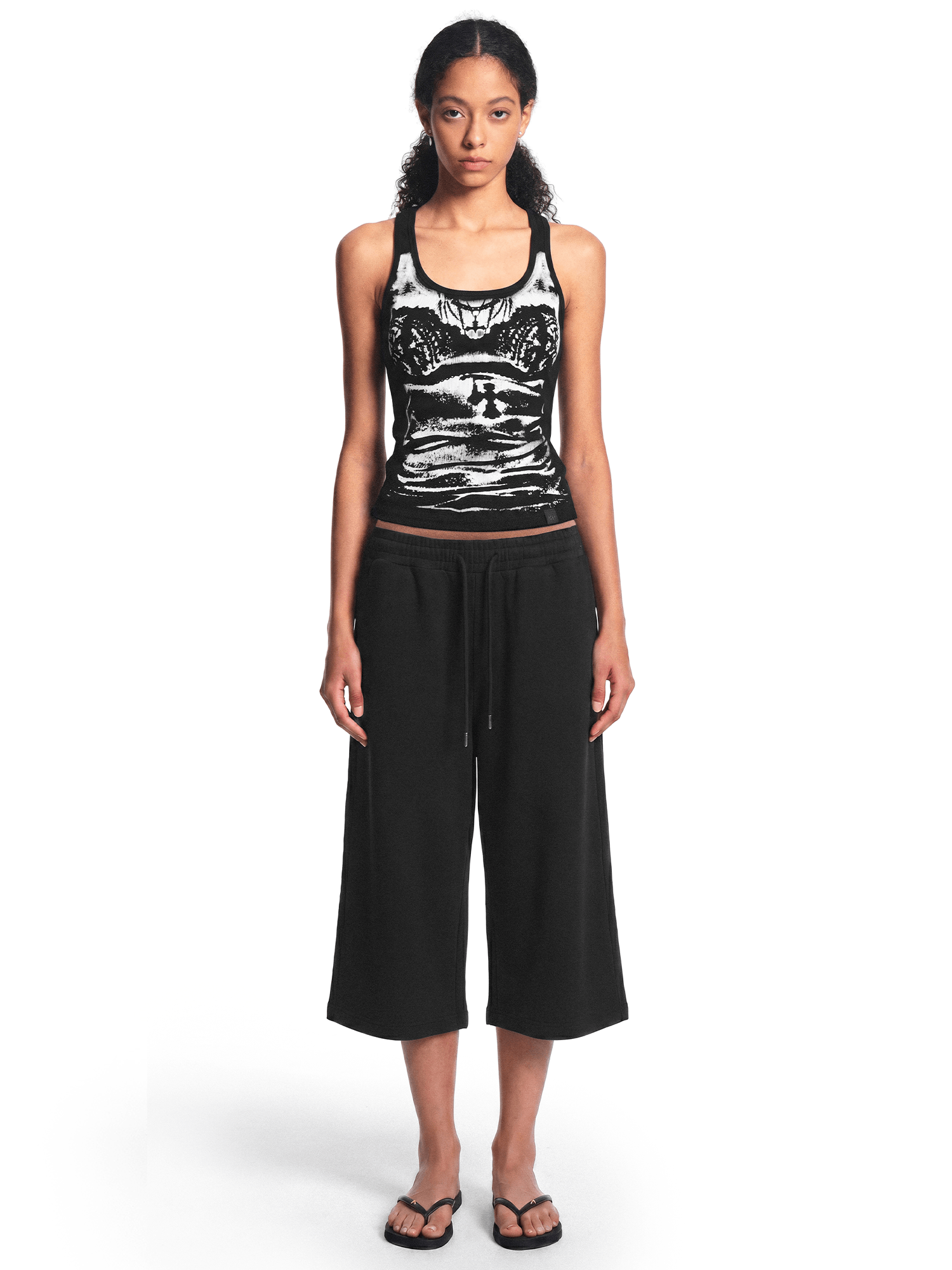 Full-body straight-on front view of a model wearing the black graphic tank top and black cropped pants with sandals.