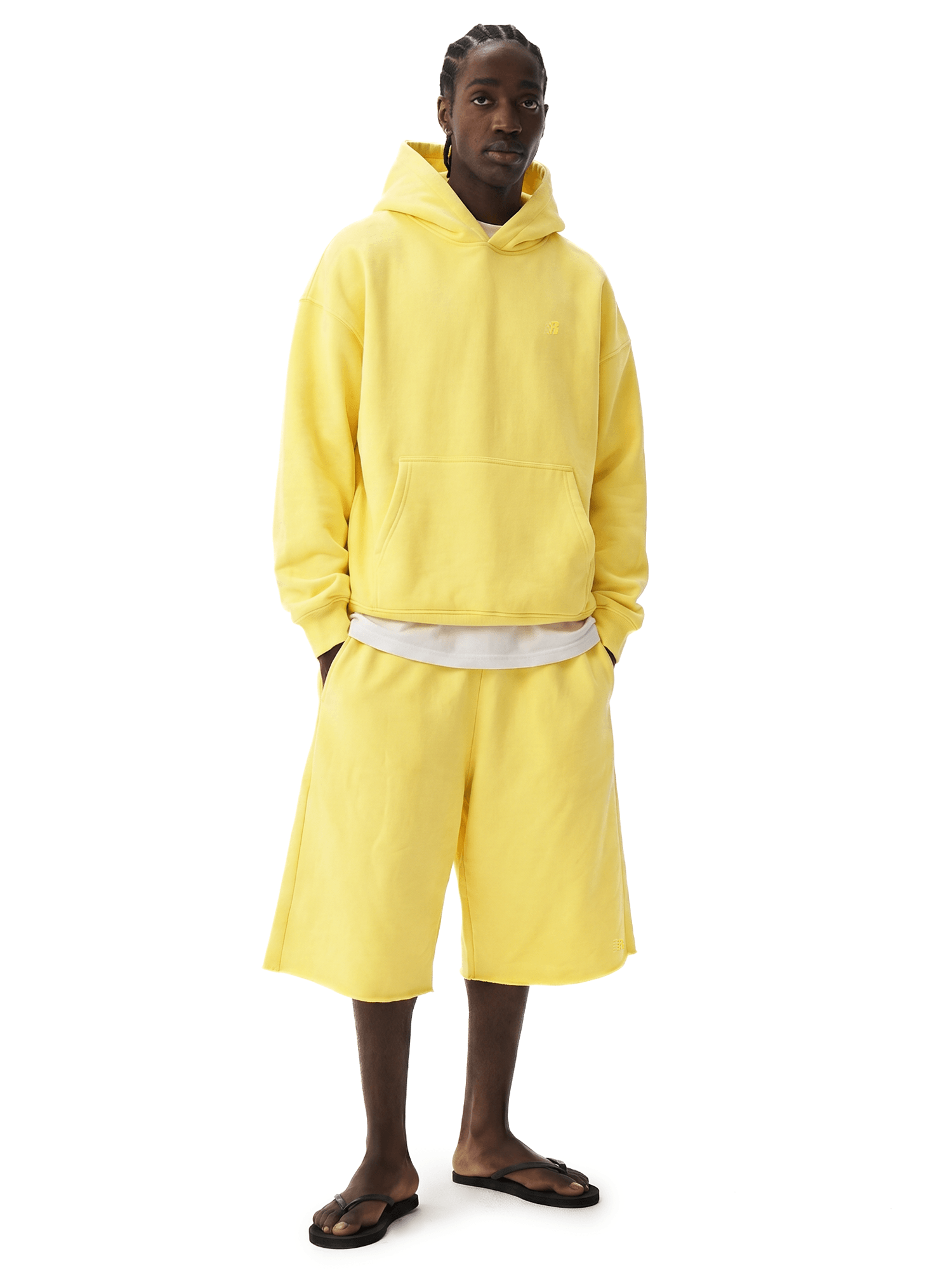 Full-body front view of a dark-skinned male model wearing a bright yellow hoodie, a light-colored undershirt, and matching bright yellow over-the-knee loose shorts, paired with flip-flops.