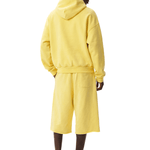 Full-body back view of a dark-skinned model wearing a bright yellow hoodie and matching loose over-the-knee shorts, looking over their shoulder slightly to the right against a plain white background.