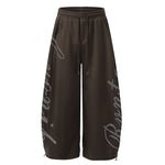 Front view of oversized dark brown wide-leg sweatpants with white script logo on the sides.