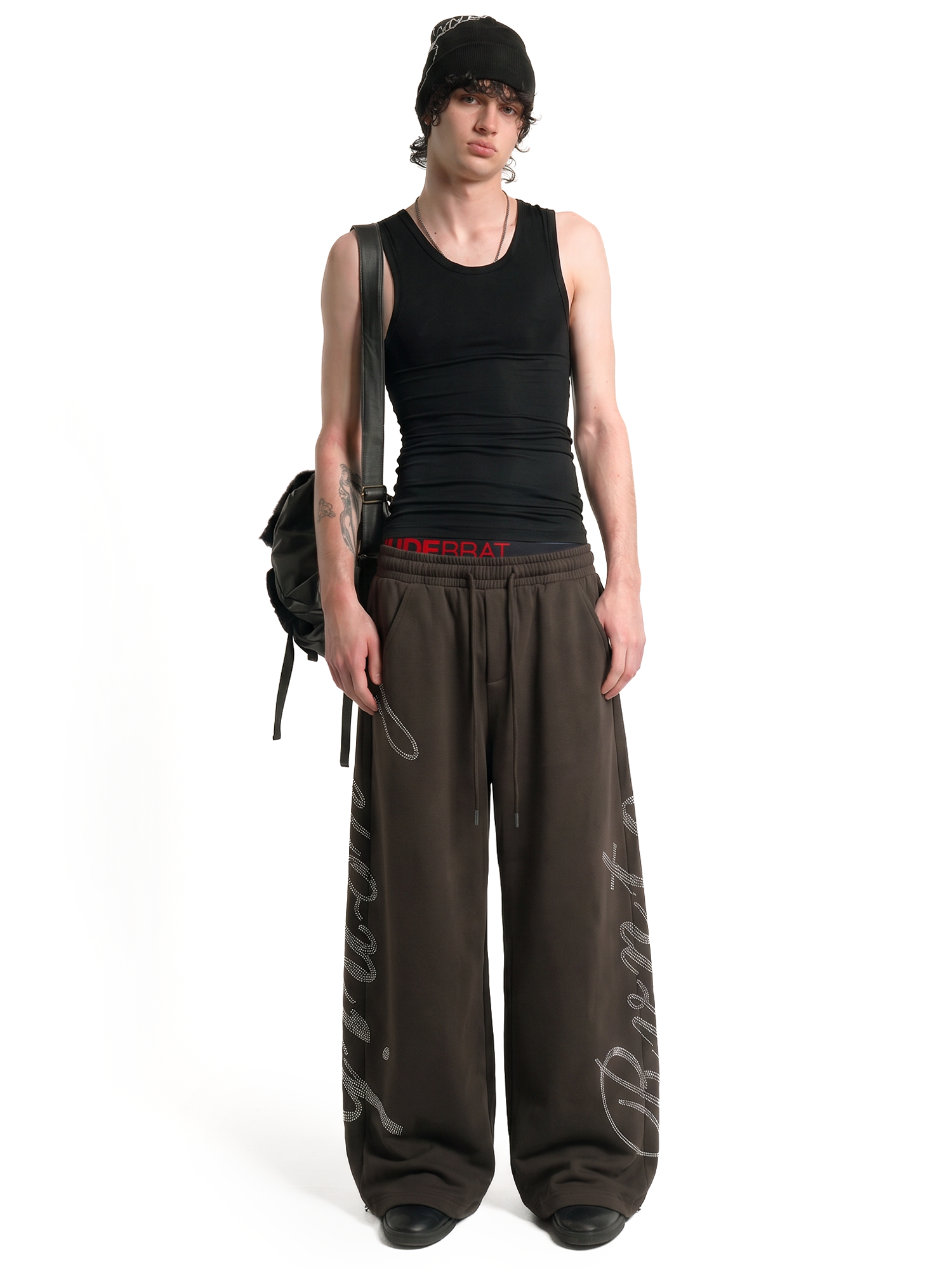 Male model wearing oversized dark brown wide-leg sweatpants with white graphic print and black tank top, street style fashion.
