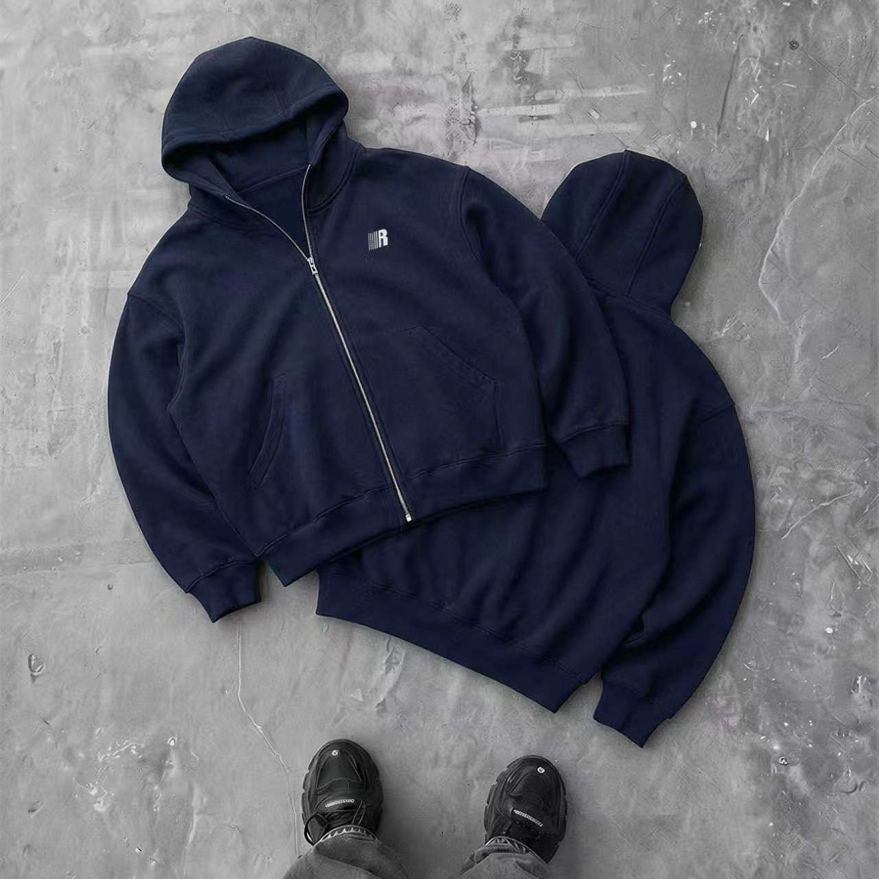 A flat-lay or top-down view of a dark navy or black hoodie styled with grey pants on a textured pavement background.