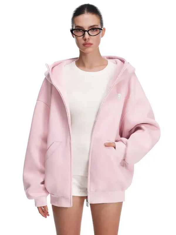Modèle en sweat à capuche rose pâle, mains dans les poches, montrant une texture de tissu lisse et une marque discrète