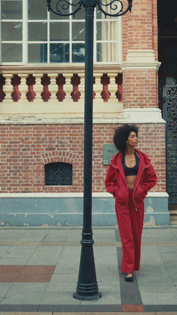 Lifestyle video of a model wearing red oversized zip-up hoodie and matching sweatpants, walking outdoors by a lake with a coffee cup, showcasing a casual streetwear style.