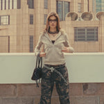 A model showcases a trendy urban look wearing a beige fur-lined hoodie, layered necklaces, and camo trousers, holding a black handbag in a city setting.