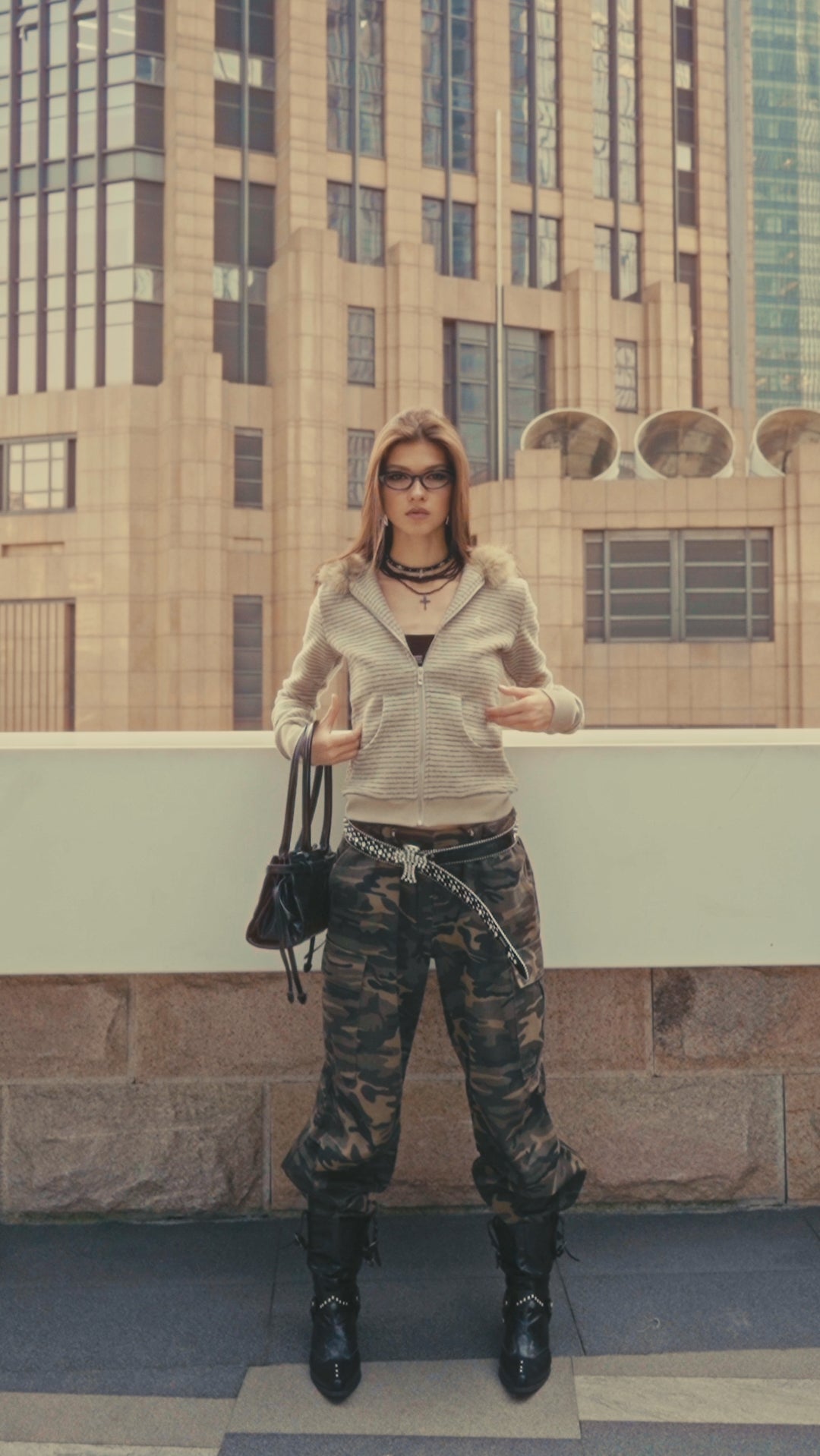 A model showcases a trendy urban look wearing a beige fur-lined hoodie, layered necklaces, and camo trousers, holding a black handbag in a city setting.