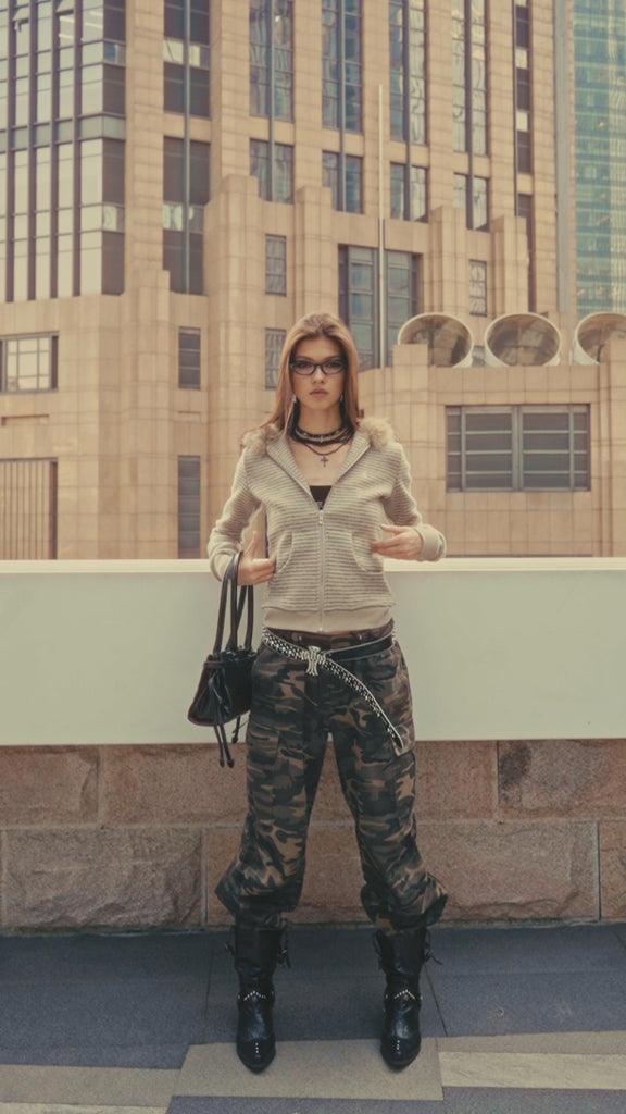 A model showcases a trendy urban look wearing a beige fur-lined hoodie, layered necklaces, and camo trousers, holding a black handbag in a city setting.